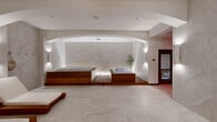 This refined spa and wellness space offers a serene, hotel-caliber retreat designed for relaxation and rejuvenation. The layout is anchored by a central wellness area showcasing a large jetted jacuzzi and a separate soaking tub, both elevated on warm teak wood platforms with subtle under-lighting that adds a luxurious glow. A graceful arched ceiling frames this focal point, lending architectural elegance to the space. The walls and floors are clad in large-format marble-effect tiles in soft, neutral tones, creating a cohesive and calming aesthetic throughout. Wall-mounted sconces provide warm, ambient lighting that complements the overall tranquil atmosphere. Adjacent to the main wellness area, a dedicated relaxation lounge accommodates multiple teak-framed chaise lounges with plush white cushions, ideal for pre- or post-treatment rest. A separate room, illuminated with a distinctive red accent light, suggests an additional treatment or sauna area. Connecting hallways guide visitors seamlessly between zones. A delicate potted olive tree introduces a natural, organic element to the interior. The overall ambiance is one of understated luxury — calm, clean, and thoughtfully designed for total well-being.