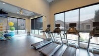 This modern fitness center is designed to support a comprehensive workout experience across two distinct zones. The cardio area is lined with large, floor-to-ceiling windows fitted with roll-down shades, flooding the space with natural light while offering open views of the surrounding community. Multiple treadmills and elliptical machines, each equipped with individual monitors, are arranged to take full advantage of the outdoor sightlines. A ceiling fan and recessed lighting keep the environment comfortable throughout.

The strength training area offers a striking contrast in character, featuring exposed brick accent walls that lend a more industrial aesthetic. Large mirrors line the walls, enhancing the sense of space while providing visual feedback during workouts. This section is equipped with a full cable machine system, a comprehensive dumbbell rack with a wide range of weights, weight benches, and additional strength machines. Wall-mounted televisions are positioned throughout for entertainment during exercise.

The flooring throughout is a durable, dark rubberized material suited for high-traffic fitness use. The soaring ceiling height adds to the open, airy ambiance of the space. Direct access to an outdoor area is available through glass-panel doors in the strength zone, seamlessly connecting the interior fitness environment with the exterior surroundings.
