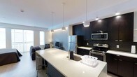 This single-floor space showcases a contemporary open-concept design that seamlessly integrates the kitchen and living area into one cohesive environment. The kitchen is a standout feature, fitted with dark cabinetry that creates a striking contrast against the bright white countertops. Stainless steel appliances, including a refrigerator, electric range, and built-in microwave, lend a polished, modern aesthetic. A large central island with a white quartz-style surface extends the workspace and doubles as a casual dining area, accommodated by sleek bar stools. Pendant lighting hangs above the island, adding both functional illumination and a design focal point against the white ceiling. The open layout flows naturally into the living area, which benefits from natural light streaming through windows along the exterior wall. The neutral tones of the living space complement the bold kitchen palette, creating a balanced and inviting atmosphere throughout. The overall design reflects a clean, minimalist sensibility with carefully chosen finishes that prioritize both style and functionality.