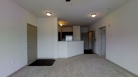 This single-floor space showcases an open-concept design that seamlessly connects the living area to the kitchen. The living area is finished with wall-to-wall carpeting, creating a comfortable and cohesive feel throughout the main space. A ceiling fan fixture is positioned overhead, contributing to the room's functionality.

The kitchen is defined by a raised peninsula with a light-toned countertop, providing both workspace and a natural visual divider between the living and cooking areas. Dark cabinetry lines the kitchen walls, contrasting with the lighter countertops and white walls. A stainless steel refrigerator is visible, and track lighting above the kitchen area ensures adequate task illumination. The entryway features hard flooring, clearly delineating the transition into the main living space.

Multiple doorways are visible throughout the space, suggesting access to additional rooms such as bedrooms or bathrooms. The walls are finished in a clean, neutral white throughout, and flush-mount ceiling lights provide consistent ambient lighting across the open layout, giving the space a practical and well-lit character.