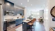 This contemporary single-floor residence showcases a thoughtfully designed open-concept layout that seamlessly connects the kitchen, dining, and living areas. The kitchen is a standout feature, outfitted with bold slate-blue cabinetry, stainless steel appliances, a marble-style backsplash, light quartz countertops, and a central island with bar seating — ideal for both cooking and casual dining. Rich hardwood-style flooring runs throughout, lending warmth and continuity to the space.

The living area is bright and airy, anchored by oversized floor-to-ceiling windows that flood the room with natural light and offer views of the surrounding greenery. A secondary sitting or reading nook shares the same generous window presence, creating an inviting retreat bathed in sunlight.

The bedroom features a striking terracotta accent wall, large windows with curtain panels, and ample space for a full furniture arrangement. Two bathrooms serve the residence: one with a glass-enclosed walk-in shower clad in marble-style tile, and a second full bath with a soaking tub and shower combo. Both bathrooms are finished with clean white vanities and hardwood-style flooring, maintaining a cohesive, modern aesthetic throughout the entire space.