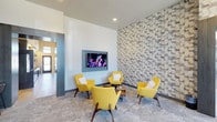 The WiFi Lounge is a thoughtfully designed communal space that blends modern aesthetics with functional comfort. The room's most striking feature is a floor-to-ceiling textured accent wall composed of interlocking geometric tiles in varying shades of grey and white, creating a dynamic, three-dimensional visual effect that anchors the space.

Centered in front of this feature wall is a curated seating arrangement of four vibrant yellow mid-century modern accent chairs, each with clean lines, tapered wooden legs, and plush cushioning. The chairs are arranged around a small central coffee table, encouraging face-to-face interaction and a relaxed atmosphere.

A wall-mounted flat-screen television is positioned at eye level, making the lounge equally suited for media viewing and casual socializing. The flooring features a neutral, patterned carpet that complements the room's cool grey and white tones while softening the space acoustically.

Recessed ceiling lighting provides even, ambient illumination throughout, and a glimpse of an adjacent corridor hints at the lounge's accessible, connected position within the broader facility. The overall ambiance is contemporary, inviting, and well-suited for both focused work and informal relaxation.