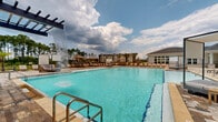 Located in Santa Rosa Beach, Florida, this single-story community amenity center offers an expansive resort-style outdoor experience designed for recreation and relaxation. The centerpiece is a generously sized swimming pool with crystal-clear water, complemented by a dramatic waterfall feature that adds both visual appeal and ambient sound. The pool deck is finished with natural stone pavers, lending a polished yet inviting aesthetic throughout the space.

A substantial wood-framed pergola anchors one end of the deck, providing partial shade and a focal point for outdoor gatherings. Surrounding the pool, rows of lounge chairs and private cabanas offer ample space for sunbathing and unwinding. An outdoor projection screen is positioned poolside, enabling open-air movie or event screenings. The covered patio area along the clubhouse exterior features ceiling fans, a ping-pong table, a wall-mounted TV, and comfortable seating arrangements, extending the usable outdoor living space.

The clubhouse itself is clad in crisp white board-and-batten siding with brick wainscoting, reflecting a coastal farmhouse architectural style. Black-framed glass doors connect the interior to the covered patio. Additional conveniences include outdoor showers, water fountains, and easily accessible restrooms. Tall Florida pines frame the perimeter, providing a natural backdrop to this well-appointed community retreat.