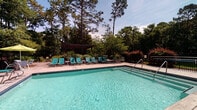 Located at 4013 Wilshire Blvd in Wilmington, NC, this single-level outdoor amenity space is thoughtfully designed for recreation and relaxation. At its centerpiece is a generously sized, crystal-clear swimming pool lined with teal lounge chairs, offering a resort-like atmosphere framed by towering pine trees and vibrant landscaping. The pool deck provides ample space for sunbathing and leisure while maintaining a serene, nature-enveloped setting.

Adjacent to the pool, a sturdy pergola structure anchors a versatile patio area finished in smooth concrete. Beneath the pergola, an outdoor ping-pong table invites casual recreation and social interaction. The patio extends to include a well-appointed outdoor kitchen featuring a stainless steel grill set into a handsome stacked-stone surround, ideal for gatherings and cookouts. Multiple seating arrangements, including white bistro-style table-and-chair sets, provide comfortable spots for dining and conversation.

A dedicated lounge area rounds out the space, furnished with Adirondack-style chairs gathered around a fire pit, creating a warm and inviting setting for evening relaxation. Lush, mature trees and manicured plantings border the entire area, providing both privacy and a verdant backdrop. The overall ambiance is one of a well-maintained, community-focused outdoor retreat designed for year-round enjoyment.