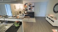 This single-floor space showcases an open-concept layout that connects the kitchen and living area in a cohesive, functional arrangement. The flooring throughout is a warm-toned hardwood-style material that adds continuity and a sense of spaciousness across the shared living spaces.

The kitchen area features dark granite or stone countertops that contrast cleanly against lighter cabinetry, with a peninsula or bar-style counter that visually separates it from the living space while maintaining an open flow. The setup offers practical workspace and a natural transition into the main living area.

The living room is comfortably scaled, centered around a dark sofa paired with a geometric-patterned area rug and a minimalist coffee table. Bold abstract artwork adorns the walls, adding color and visual interest to the otherwise neutral palette. Warm ambient lighting enhances the inviting atmosphere of the room.

A white entry door is visible at the far end of the living space, offering a clean, straightforward entry point. The overall ambiance of the space is modern and welcoming, with a neutral base accented by warm tones and contemporary décor choices throughout.