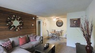 This single-floor residential space offers a comfortable and functional layout centered around an open-concept living and dining area. A standout architectural feature is the striking horizontal wood plank accent wall that adds warmth and a rustic character to the living room. The living area is carpeted and furnished with a plush sectional sofa accented with decorative pillows, complemented by a dark coffee table. Adjacent to the living space, the dining area accommodates a round wooden table with ornate metal chairs, ideal for everyday meals. A ceiling fan with a light fixture provides both ventilation and ambient lighting throughout the combined space. The walls are painted in a neutral tone, creating a clean and versatile backdrop for décor. Wall-mounted decorative accents, including a large round mirror and framed artwork, add personality to the space. A textured popcorn ceiling is present throughout. A doorway visible from the living area provides access to additional rooms, suggesting a connected and practical floor plan. Overall, the space presents a warm, lived-in ambiance with a blend of rustic and casual design elements.