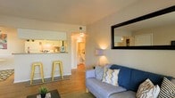 This single-floor space offers a thoughtfully designed open layout that maximizes functionality and flow. The living area serves as the central hub, featuring warm hardwood flooring, neutral-toned walls, and a large framed mirror that enhances the sense of space by reflecting natural and ambient light throughout the room. A comfortable seating area anchors the space, complemented by warm accent lighting from a floor lamp that contributes to the inviting atmosphere.

A defining architectural feature is the kitchen pass-through window, which connects the living area to the kitchen seamlessly. The pass-through is paired with a breakfast bar and metal bar stools, creating a casual dining option that encourages interaction between the kitchen and living spaces. The kitchen itself is well-lit with overhead lighting and features white cabinetry, providing ample storage.

A hallway visible from the living area leads to additional rooms, including what appears to be a bathroom with white fixtures and continued warm lighting. The consistent use of hardwood flooring and neutral color palette throughout creates a cohesive, welcoming environment that feels both practical and comfortable.