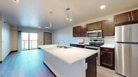 This single-floor residence offers a thoughtfully designed layout with modern finishes and functional living spaces throughout. The heart of the home is an open-concept kitchen anchored by a large center island with a built-in sink and white countertop surface, complemented by rich dark-toned cabinetry and a full suite of stainless steel appliances, including a range, microwave, and refrigerator. Pendant lighting above the island adds a polished touch, while wood-look flooring ties the kitchen and adjacent living area together seamlessly. A sliding glass door leads to a private balcony, filling the living space with natural light and offering outdoor views. The bedrooms feature plush carpeting, generous proportions, and ample natural light from large windows. One bedroom includes a walk-in closet outfitted with wire shelving systems for organized storage. The bathroom is finished with a dark wood vanity topped with a speckled countertop, a tub-shower combination, and wood-look flooring. A dedicated laundry area houses a stacked washer and dryer unit, adding everyday convenience. Throughout the residence, dark baseboards provide a clean contrast to the light-toned walls, contributing to a cohesive and contemporary aesthetic.