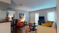 Located at 8000 Oakdell Way in San Antonio, Texas, this single-floor residence offers a thoughtfully arranged layout that balances comfort and functionality. The living room serves as the heart of the home, featuring rich hardwood-style flooring, multiple windows with slatted blinds that welcome natural light, and enough space to accommodate a full seating arrangement centered around a glass-top coffee table. The open flow connects seamlessly to a dedicated dining area, illuminated by an elegant pendant chandelier and accented by a large framed mirror that enhances the sense of space. A breakfast bar with seating bridges the dining area to the kitchen, which is equipped with warm wood cabinetry, dark granite-style countertops, and white appliances including a range and full-size refrigerator. The bedrooms feature plush carpeting, ceiling fans, and windows dressed with bold curtain panels, creating a cozy and restful atmosphere. The bathroom is finished with wood-tone cabinetry, a dark countertop vanity, a framed mirror, and a shower-tub combination. Throughout the residence, neutral wall tones provide a clean, versatile backdrop, while consistent flooring and cohesive design details contribute to an inviting and well-connected living environment.