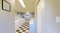 This communal laundry facility at 300 Hickory Ridge Cir in Hopkinsville, Kentucky, is a compact, efficiently organized space designed to serve residents with convenience and ease. The room is lined on both sides with rows of white top-loading washers and front-loading dryers, maximizing the available floor area and providing ample capacity for multiple users simultaneously. The machines are equipped with modern CSC app-based payment technology, allowing for a seamless and cashless laundry experience.

The interior features a clean, neutral color palette with beige walls on one end and a calming blue accent wall on the other, adorned with a decorative "Laundry" sign that adds a welcoming touch. The striking blue, black, and white checkered tile flooring runs throughout, giving the space a classic yet practical aesthetic. Overhead, flush-mounted fluorescent lighting ensures the room is well-lit at all times.

A small entrance area leads into the main laundry room, where a window allows for natural light and ventilation. A folding counter with a granite-style surface provides a functional space for sorting and handling laundry. Safety is addressed with a prominently mounted fire extinguisher. Posted service instructions ensure users have all the information needed for a smooth experience.
