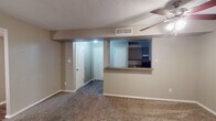 This single-story unit is designed with a practical, open layout that connects the main living and kitchen areas seamlessly. The spacious living room features wall-to-wall carpeting, a ceiling fan with light fixture, and a textured ceiling. A structural beam overhead defines the boundary between the living space and the kitchen, which is accessible via a pass-through counter with an overhang — ideal for casual dining or meal prep. The kitchen is equipped with dark appliances, including a black range, refrigerator, and dishwasher, complemented by gray-toned cabinetry and laminate countertops. Dark hardwood-style flooring runs through the kitchen, creating a contrast to the carpeted areas. Two bedrooms offer comfortable proportions, plush carpeting, ceiling fans, and window blinds providing natural light. The hallway connects both bedrooms to two bathrooms, each featuring warm wood-toned vanity cabinetry, single-basin sinks with mirrored medicine cabinets, and a tub-shower combination. One bathroom includes a separate textured-wall entry and wood-style flooring. Neutral beige tones throughout create a cohesive, understated ambiance.