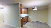 This single-floor residence offers a practical and comfortable layout designed for everyday living. The kitchen is a standout feature, showcasing warm medium-toned wood cabinetry that extends from counter height to the ceiling, providing generous storage space. Dark countertops offer a grounding contrast to the cabinetry, while an undermount sink is neatly positioned beneath upper cabinets with integrated lighting. A peninsula-style counter extends into the space, creating a natural division between the kitchen and the adjacent living area. A stylish pendant light with a brushed metal finish hangs above, adding a decorative focal point to the transition zone between rooms. The living area is finished with plush carpeting, contributing to a cozy and inviting atmosphere. A baseboard heating unit runs along the wall, ensuring efficient warmth throughout the space. The textured ceiling adds a classic touch, while neutral wall tones keep the overall palette light and versatile. The open-concept flow between the kitchen and living area makes the space feel connected and well-suited for both everyday use and casual entertaining.