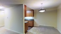 This single-floor residence offers a practical and comfortable layout designed for everyday living. The kitchen is a standout feature, showcasing warm medium-toned wood cabinetry that extends from counter height to the ceiling, providing generous storage space. Dark countertops offer a grounding contrast to the cabinetry, while an undermount sink is neatly positioned beneath upper cabinets with integrated lighting. A peninsula-style counter extends into the space, creating a natural division between the kitchen and the adjacent living area. A stylish pendant light with a brushed metal finish hangs above, adding a decorative focal point to the transition zone between rooms. The living area is finished with plush carpeting, contributing to a cozy and inviting atmosphere. A baseboard heating unit runs along the wall, ensuring efficient warmth throughout the space. The textured ceiling adds a classic touch, while neutral wall tones keep the overall palette light and versatile. The open-concept flow between the kitchen and living area makes the space feel connected and well-suited for both everyday use and casual entertaining.
