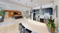 Located at 743 Montague Expy in Milpitas, California, this single-floor community amenity space is thoughtfully designed to offer both comfort and functionality. The layout flows seamlessly from one zone to the next, all finished with light wood-tone flooring that unifies the space with a clean, contemporary aesthetic.

The lounge area makes an immediate impression with a bold orange accent wall, a large flat-screen television, and stylish furnishings including a plush sofa, cushioned armchairs, and a gold-accented coffee table atop a patterned area rug. A decorative pendant chandelier and geometric wall art add artistic flair to the space.

Adjacent to the lounge is a well-appointed kitchen featuring dark cabinetry, stainless steel appliances including a full-size refrigerator and microwave, and a generous white quartz island with bar seating beneath striking teardrop pendant lights. The kitchen is designed for communal use and social gatherings.

A conference and collaboration zone offers a long communal table with ample seating, anchored by a large wall-mounted display — ideal for meetings or group activities. Recessed lighting throughout ensures consistent illumination across all areas.

Large glass doors open to an outdoor patio with seating, bringing in natural light and views of the surrounding property. Potted plants and curated décor elements add warmth and a welcoming ambiance throughout.