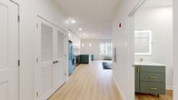 This thoughtfully designed single-floor unit offers a bright, open-concept layout that seamlessly connects the kitchen, dining, and living areas. Light hardwood flooring flows throughout the entire space, complementing the crisp white walls and recessed lighting that keep the interior feeling airy and inviting.

The kitchen is a standout feature, showcasing a two-tone cabinetry design with white upper cabinets and sage green lower cabinets, both accented with gold hardware. A marble-style backsplash adds an elegant touch, while the extended peninsula with a white countertop provides both prep space and casual seating. Stainless steel appliances, including a gas range, microwave, and dishwasher, are joined by a retro-style blue refrigerator that adds a distinctive design flair. Pendant lighting above the peninsula enhances the kitchen's modern aesthetic.

The living area is generously proportioned, offering ample room for comfortable furnishings, and benefits from natural light via a window overlooking the outdoors. A louvered closet door provides convenient storage nearby.

The bathroom features a floating sage green vanity with gold hardware, a white countertop, and an illuminated LED mirror, maintaining the cohesive color palette found throughout the unit. The space is well-appointed with clean, contemporary finishes at every turn.