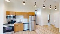 Located at 204 High St in Buffalo, NY, this one-bedroom, one-bathroom unit blends historic character with modern finishes across a single, efficiently designed floor. The open kitchen anchors the living space and is outfitted with warm-toned wood cabinetry, stainless steel appliances including a range, microwave, dishwasher, and refrigerator, plus light countertops and a single-basin sink. Stylish pendant lighting hangs from notably tall ceilings, enhancing the sense of openness throughout. Exposed brick walls appear prominently in the kitchen, living area, bedroom, and bathroom, lending an industrial, loft-like character that contrasts beautifully with the clean white walls and light hardwood-style flooring found throughout the unit. The bedroom is comfortably sized and features a built-in closet with shelving and a bright window overlooking the surrounding neighborhood. The bathroom continues the brick aesthetic with a large window that fills the space with natural light. A hallway connects the rooms, providing a logical flow. Overall, the unit offers a cohesive blend of historic architectural detail and contemporary updates.