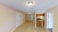 This single-floor space presents a practical, open layout that connects the main living area to the kitchen. The living and entry area features light beige tile flooring throughout, contributing to a clean, cohesive look. Walls are painted in a neutral tone, complemented by white trim and doors, creating a bright and airy atmosphere. Flush-mount ceiling light fixtures provide consistent overhead lighting, and a textured popcorn ceiling adds a classic touch.

The kitchen, visible from the main living area and partially separated by a half-wall with a countertop overhang, features warm wood-toned cabinetry and a light countertop. Under-cabinet lighting illuminates the workspace, and the layout allows for easy interaction between the kitchen and the adjacent living space.

Two white panel doors are positioned near the entry, likely leading to a closet and an additional room or utility space. A fire extinguisher is mounted on the wall, highlighting attention to safety. A doorway at the far end of the hallway leads further into the unit, suggesting additional rooms beyond the main living and kitchen area. Overall, the space offers a functional and welcoming environment.