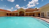 Located at 9000 Colby Dr in Fort Myers, Florida, this single-story community amenity center showcases a warm Mediterranean-inspired design, highlighted by a distinctive red tile roof, stucco exterior in soft golden hues, and elegant arched architectural details throughout. At the heart of the facility is a generously sized outdoor swimming pool with a wide paver surround, offering ample space for lounging under the Florida sun, framed by lush tropical greenery and palm trees that enhance the resort-like atmosphere. Adjacent to the pool, a spacious covered patio area features decorative columns, exposed beam ceilings, and ceiling fans, creating a comfortable shaded retreat furnished with contemporary seating arrangements including sofas, lounge chairs, and dining sets — ideal for relaxing or socializing. The tile-floored pavilion also serves as a recreational hub, equipped with yard games such as cornhole and a giant Connect Four, providing entertainment options for residents of all ages. A set of stately double entry doors with arched transom windows anchor the main building entrance, adding a refined architectural focal point. The overall ambiance blends casual Florida outdoor living with classic design elements, making this facility a welcoming and functional community gathering space.