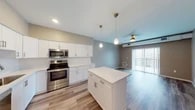 This well-appointed single-floor unit offers a thoughtfully designed open-concept layout that seamlessly connects the kitchen and living areas. The kitchen is a standout feature, showcasing crisp white shaker-style cabinetry, a full suite of stainless steel appliances including a range, over-the-range microwave, and refrigerator, as well as a white quartz countertop that extends to a central island. Elegant teardrop pendant lights hang above the island, complementing the recessed ceiling lighting and adding a refined touch to the space. Light wood-look flooring runs throughout the main living and kitchen areas, contributing to a bright, contemporary feel. The living area benefits from a ceiling fan and large sliding glass doors that invite natural light and provide access to an outdoor balcony. The bedroom is finished with soft carpeting, a ceiling fan, and a window offering natural light and exterior views. The bathroom presents a clean, minimalist aesthetic with white cabinetry, a white quartz vanity, an undermount sink, a large wall mirror, and a glass-enclosed shower with tile surround. Neutral wall tones throughout create a cohesive and welcoming ambiance across the entire unit.