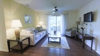 This single-floor space showcases a thoughtfully designed living area that combines comfort and functionality. The living room features rich, dark hardwood flooring that anchors the space and adds warmth throughout. Soft yellow-green walls create an inviting, airy atmosphere, complementing the abundance of natural light streaming through a large sliding glass door fitted with sheer white curtains. The door provides both an outdoor view and convenient access to an exterior area, enhancing the sense of openness.

A ceiling fan with a classic dark finish is centrally positioned overhead, offering both practical airflow and a subtle decorative touch. The seating arrangement includes a spacious sofa adorned with coordinating throw pillows, paired with a striped armchair, creating a balanced and cohesive conversation area. A glass-topped coffee table sits atop a patterned area rug, adding texture and visual interest to the hardwood floor beneath. Ambient table lamps on either side of the room provide warm, layered lighting for the evenings.

Overall, the space presents a clean, comfortable, and well-lit environment with a neutral yet cheerful color palette that feels both practical and welcoming.
