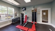 Located at 10017 Season Grove Ln in Charlotte, NC, this single-floor fitness center is a well-equipped exercise facility designed to support a full range of workout routines. The space features soft sage-green walls, durable rubber flooring, and bright flush-panel ceiling lighting supplemented by a ceiling fan, creating a clean and comfortable environment for exercise.

The cardio area includes multiple treadmills and elliptical machines positioned near large, multi-pane windows that allow ample natural light and provide views of the surrounding green landscaping, keeping the space feeling open and connected to the outdoors.

The strength training area is thoughtfully arranged and includes a multi-station cable machine with lat pulldown and leg extension attachments, a leg curl/extension bench, adjustable workout benches with red upholstery, and a well-stocked dumbbell rack. A wall-mounted mirror expands the visual depth of the room and allows users to monitor their form during workouts.

A wall-mounted television is positioned for visibility across the strength training zone, adding an entertainment option during workouts. Safety is addressed through clearly marked exit signage and door alarms. The facility offers a practical, fully functional workout environment suitable for both cardio and resistance training needs.