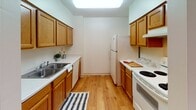 Located at 1115 Second St NE in Minneapolis, Minnesota, this single-floor unit offers a practical and functional layout with several notable architectural details. The space is organized to maximize efficiency while providing comfortable living areas throughout.

The kitchen follows a galley-style configuration, featuring warm honey-toned wood cabinetry lining both walls, white appliances including a refrigerator, electric range, and dishwasher, and a double basin sink. Light hardwood flooring runs the length of the kitchen, and a large overhead skylight panel provides bright, even illumination.

Carpeted throughout the remaining living areas, the space transitions into a generously proportioned main room with a vaulted ceiling accented by exposed wooden beams and a ceiling fan. Multiple windows allow natural light to filter in, and baseboard heating units run along the walls. A second carpeted room, accessible via a dark-framed door, features a wall-mounted air conditioning unit and a pendant light fixture.

A hallway connects the rooms and leads to the bathroom, which includes a vanity with oak cabinetry, a single-basin sink, a large mirror with overhead lighting, and a shower/tub combination with a curtain. A spacious walk-in closet with wire shelving provides substantial storage. Dark wood door frames appear consistently throughout, adding visual contrast to the neutral wall tones.
