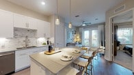 This well-appointed single-floor residence showcases a thoughtfully designed open-concept layout with modern finishes throughout. The kitchen is a standout feature, outfitted with white shaker cabinetry, stainless steel appliances, a marble-style tile backsplash, under-cabinet lighting, and a generous island with seating — ideal for both cooking and casual dining. Pendant lighting above the island adds a refined touch, while light hardwood-style flooring extends seamlessly into the living area. The living room is bright and inviting, anchored by French doors that open to an outdoor view, allowing abundant natural light to fill the space. A ceiling fan, curated wall art, and a cozy area rug contribute to the room's relaxed yet polished ambiance. The bedrooms offer a serene retreat, featuring plush carpeting, large windows dressed with sheer curtains and blinds that frame greenery outside, and neutral tones that promote restful comfort. The primary bathroom includes a glass-enclosed walk-in shower with marble-style tile surround, a white dual-sink vanity with gray countertops, and wood-look flooring. A secondary bathroom provides a tub-shower combination and a matching white vanity. Together, these spaces create a cohesive, contemporary living environment with quality details at every turn.