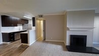 This single-floor residence offers a functional and comfortable open-concept layout that seamlessly connects the main living area with the kitchen. The living room is finished with plush wall-to-wall carpeting and features a classic fireplace with a white-painted mantel and surround, serving as a natural focal point of the space. Crown molding runs along the ceiling, adding a refined architectural detail throughout the main area. The textured ceiling and neutral beige walls create a warm, understated ambiance. The kitchen is positioned adjacent to the living space and is separated by a partial wall counter, offering both an open feel and a defined cooking zone. Dark espresso-toned cabinetry lines the kitchen walls, complemented by under-cabinet lighting and a stainless steel refrigerator. A hallway visible from the living area leads to additional rooms, suggesting a practical and well-organized floor plan. The space is efficiently laid out, making good use of the available footprint while maintaining a sense of openness between the key living and cooking areas.