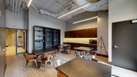 This single-floor space showcases a striking industrial-modern aesthetic, characterized by soaring ceilings with exposed ductwork and piping, linear pendant lighting, and a neutral gray and dark color palette. The open layout is organized around multiple dark-topped tables paired with polished metal and wood bistro-style chairs, offering flexible seating arrangements well-suited for collaborative work, events, or casual gatherings.

Along one wall, a run of rich dark wood cabinetry with under-cabinet lighting provides ample storage, while a large open steel shelving unit displays an assortment of supplies and accessories. An artist's easel and potted greenery add a creative, welcoming touch to the space.

A bold dark charcoal accent wall anchors one end of the room, featuring a door that leads to an adjacent area. On the opposite end, a wall of tall, narrow windows floods the space with natural light and offers views of an outdoor terrace with landscaping, framing the exterior environment beautifully.

Light wood-tone flooring runs throughout, softening the industrial elements and creating a warm, inviting atmosphere. Decorative artwork, including celestial-themed prints, adds personality and character to the walls. The overall ambiance is contemporary and versatile, blending functionality with thoughtful design details.