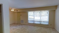 This single-story space presents a straightforward and functional layout centered around a spacious main living area. The room is finished with wall-to-wall carpeting in a neutral beige tone, contributing to a quiet and understated aesthetic throughout. A prominent bank of large windows along one wall allows a generous amount of natural light to fill the room, fitted with horizontal blinds that offer adjustable privacy and light control. The windows provide views of surrounding greenery, adding a pleasant outdoor connection to the interior. The walls are painted in a soft, warm neutral tone that complements the carpeted flooring. A ceiling fan is installed overhead, supporting air circulation throughout the space. A floor-level HVAC vent is positioned along the lower wall near the windows, indicating a built-in climate control system. A standard interior door is visible in the corner of the room, suggesting access to an adjacent area or utility space. The overall design is clean and minimal, offering a blank-canvas environment that is well-suited to a variety of furniture arrangements and uses.