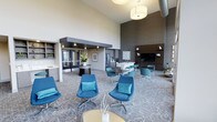 This single-floor space is a thoughtfully designed communal lounge that blends modern aesthetics with functional comfort. The room immediately impresses with its soaring ceilings, which lend an open, airy atmosphere and amplify the sense of scale throughout the interior. Exposed industrial-style ductwork contrasts elegantly with the smooth white walls and warm pendant lighting overhead.

The seating arrangement centers around a collection of vibrant teal lounge chairs and accent chairs, each accented with coordinating cushions. These are organized into both intimate conversation clusters and small dining groupings with sleek dark tables, catering to a variety of social settings. The patterned carpet in soft gray tones adds visual texture underfoot while tying the design together cohesively.

A dark, textured feature wall anchors one end of the room, housing a large flat-screen television and built-in cabinetry with decorative figurines on display. Adjacent to this, a compact kitchenette with a light countertop and stainless steel appliances provides added convenience. Near the entrance, glass-paneled double doors welcome natural light, while open shelving and a display counter offer additional functionality. The overall ambiance is sleek, welcoming, and well-appointed.