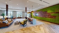 Located at 2225 Collett Ave in Corona, California, this single-floor community clubhouse showcases a boldly designed interior that blends modern aesthetics with natural, organic elements. The entrance immediately sets the tone with striking blue-and-white herringbone tilework lining an accent wall, complemented by a rich wood credenza adorned with hanging potted plants and vibrant botanical artwork.

The open-plan layout features wide-plank hardwood floors laid in a classic herringbone pattern, flowing seamlessly across the space and lending warmth and visual continuity throughout. Generous ceiling heights and abundant recessed lighting create a bright, airy atmosphere, further enhanced by large, grid-paned windows that invite natural light and offer outdoor views.

A central lounge area is furnished with mid-century modern seating, including a leather sofa and accent chairs arranged atop a layered area rug. Nearby, a long communal high-top table surrounded by cognac leather bar stools is suspended beneath oversized gold dome pendant lights, forming an ideal collaborative or social workspace.

One of the space's most distinctive features is a suspended rope swing sofa framed by floor-to-ceiling industrial-style windows and a dramatic living moss wall paired with a textured wood-and-stone accent wall housing a flat-screen display. A sculptural twig chandelier and circular wall art add artistic flair. The overall ambiance is energetic, welcoming, and thoughtfully curated.