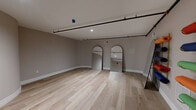This 310 square foot studio space showcases a blend of modern design and industrial elements. The room's most striking feature is its curved walls, creating a unique, flowing layout. Light wood flooring spans the entire space, adding warmth and continuity. Large arched mirrors are strategically placed, enhancing the sense of space and light. 

The studio's design incorporates exposed black piping along the ceiling, contributing to an industrial aesthetic. This is complemented by recessed lighting fixtures that provide even illumination throughout. A standout decorative element is the set of colorful, cylindrical objects mounted on the wall, adding a playful pop of color to the neutral palette.

The space includes a door with a narrow window, likely leading to a hallway or exterior. White baseboards and trim provide a clean finish to the walls. The room's layout and features suggest it could serve various purposes, from a creative workspace to a compact living area, offering flexibility in its use.