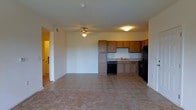 This single-floor unit presents an open-concept layout where the living area and kitchen flow seamlessly together, creating a functional and cohesive space. The living area is finished with wall-to-wall carpeting in a neutral tone, providing a comfortable foundation for furnishings. A ceiling fan with a built-in light fixture is centrally positioned overhead, offering both air circulation and ambient lighting throughout the combined space.

The kitchen is situated along the back wall and features warm, medium-toned wood cabinetry that extends both above and below the countertops, providing ample storage. A black dishwasher, black refrigerator, and a sink with under-cabinet lighting make up the kitchen's core appliances and fixtures. The kitchen floor transitions to tile, clearly delineating the cooking area from the carpeted living space.

The unit's neutral white walls and standard-height ceilings contribute to a clean, open feel. A hallway visible on the left side of the space leads to additional rooms, and the overall layout suggests an efficient and practical floor plan suited for everyday living.