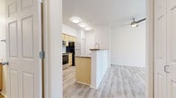 This single-floor residence offers a clean, functional layout with light wood-style flooring running consistently throughout the space, creating a cohesive and airy atmosphere. The kitchen is designed as a galley-style space, featuring light wood cabinetry, dark stone countertops, a double-basin sink with a modern gooseneck faucet, and stainless steel appliances including a range, microwave, refrigerator, and dishwasher. A peninsula with matching cabinetry separates the kitchen from the living area, providing a natural transition between the two spaces. The living area is bright and open, benefiting from a large window and a sliding glass door fitted with vertical blinds that lead to a private outdoor patio, filling the room with natural light and outdoor views. A ceiling fan adds comfort to the space. The bedroom areas feature the same light flooring, with one room offering built-in upper cabinetry and a built-in desk shelf — an ideal workspace or study nook. The hallway connects seamlessly to a bathroom equipped with a full tub and shower surround, chrome fixtures, and a towel bar. A vanity with warm wood cabinetry and a well-lit mirror completes the bathroom. The overall palette of whites and natural wood tones gives the space a fresh, welcoming feel.