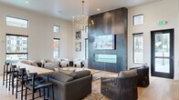This thoughtfully designed single-floor communal space blends modern sophistication with inviting comfort across a generously proportioned open floor plan. The living area commands attention with soaring ceilings, floor-to-ceiling dark metal accent panels housing a linear glass fireplace and a mounted flat-screen TV, and a bold brass candelabra-style chandelier overhead. Large black-framed windows and clerestory windows flood the space with natural light while offering views of the outdoor pool and patio area. Multiple seating arrangements — including a large sectional sofa, lounge chairs, and intimate booth-style nooks — create a versatile social environment. Deep forest green accent walls with architectural arch detailing add warmth and character throughout. The kitchen area is equally impressive, featuring rich dark cabinetry with brass hardware, a substantial waterfall-edge marble island with built-in seating, and a suite of pendant lighting in varied contemporary styles. Light hardwood flooring unifies the space seamlessly. A game area with a pool table, communal dining tables, and double glass-panel entry doors leading directly to the outdoor spa and pool complete this well-appointed amenity space.