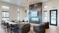 This thoughtfully designed single-floor communal space blends modern sophistication with inviting comfort across a generously proportioned open floor plan. The living area commands attention with soaring ceilings, floor-to-ceiling dark metal accent panels housing a linear glass fireplace and a mounted flat-screen TV, and a bold brass candelabra-style chandelier overhead. Large black-framed windows and clerestory windows flood the space with natural light while offering views of the outdoor pool and patio area. Multiple seating arrangements — including a large sectional sofa, lounge chairs, and intimate booth-style nooks — create a versatile social environment. Deep forest green accent walls with architectural arch detailing add warmth and character throughout. The kitchen area is equally impressive, featuring rich dark cabinetry with brass hardware, a substantial waterfall-edge marble island with built-in seating, and a suite of pendant lighting in varied contemporary styles. Light hardwood flooring unifies the space seamlessly. A game area with a pool table, communal dining tables, and double glass-panel entry doors leading directly to the outdoor spa and pool complete this well-appointed amenity space.