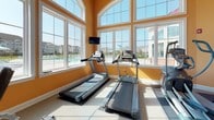 Located at 100 Isabelle Isle in Dover, Delaware, this fitness center offers a well-equipped, single-floor exercise facility designed to support a comprehensive workout experience. The space is immediately distinguished by its warm golden-orange walls and an abundance of natural light, courtesy of large arched and floor-to-ceiling windows that frame views of the surrounding outdoor pool area and community grounds.

The main exercise floor is generously sized, providing ample room for a varied selection of cardio and strength-training equipment. Treadmills are positioned directly in front of the expansive window wall, allowing users to enjoy bright, open views while working out. Additional cardio options include an elliptical trainer and a rowing machine, complemented by a multi-function cable strength station suited for a full-body workout. A wall-mounted television adds convenience for users during their sessions.

A pass-through mirror window connects the main workout area to an adjacent room, which features bold blue accent walls and a water fountain, suggesting a transitional or stretching space. Recessed lighting and carpeted flooring in sections of the facility contribute to a clean, comfortable environment. The overall ambiance is energetic yet welcoming, combining ample sunlight, community views, and a practical equipment layout to create an inviting space for daily fitness routines.