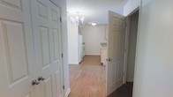 This single-floor space is characterized by its clean, neutral aesthetic and practical layout. The entryway opens into a central hallway with light hardwood-style flooring that extends throughout the main living areas, lending warmth and visual continuity to the space.

The kitchen is positioned at the far end and features white cabinetry, white appliances including a refrigerator and range, and light countertops. The layout is straightforward and functional, offering ample cabinet storage for everyday use. Adjacent cabinetry near a secondary doorway suggests additional storage or utility space nearby.

Overhead, a decorative chandelier with candle-style lights serves as a focal point in the dining or transition area, adding a touch of elegance to the otherwise understated interior. A flush-mount ceiling fixture provides supplementary lighting in the kitchen area.

The walls are finished in a soft white tone throughout, contributing to a bright and open feel. White six-panel doors are consistent across the space, reinforcing the clean, uniform design. The textured ceiling adds a subtle traditional character to the overall interior.