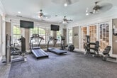 Located at 100 Parkway Cir S in Atlanta, GA, this well-appointed fitness center offers a comprehensive workout environment within a thoughtfully designed single-floor space. The room features warm taupe walls accented by crisp white crown molding and baseboards, lending an upscale feel uncommon in typical gym facilities. Large windows along one wall flood the space with natural light and provide direct views of an outdoor pool area, creating an inviting and motivating atmosphere. Elegant French doors serve as the primary entrance, opening to a stairwell and adding a classic architectural touch.

The cardio section is equipped with multiple treadmills, an elliptical trainer, and a stationary bike, each positioned near wall-mounted flat-screen televisions for entertainment during workouts. The strength training area includes a multi-function cable machine, a weight bench, a dumbbell rack, and additional resistance equipment. An adjacent storage nook provides space for exercise accessories such as resistance bands and stability balls.

Durable, dark carpet tiles cover the floor throughout, offering both comfort and practicality. A ceiling fan with recessed lighting ensures proper ventilation and illumination. Sanitation supplies and a wall-mounted dispenser are conveniently positioned near the entrance, reflecting attention to cleanliness and user convenience. This fitness center delivers a complete workout experience within a refined and well-maintained setting.