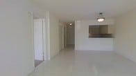 This single-floor space features a bright, open layout with a clean and minimalist aesthetic throughout. The main living area is finished with large white ceramic tile flooring that extends seamlessly across the space, reflecting light and contributing to an airy, spacious feel. Walls are painted in crisp white, maintaining a neutral palette that enhances the sense of openness.

A pass-through window connects the living area to the kitchen, which features medium-toned cabinetry and a dark countertop. The kitchen layout appears compact yet functional, with overhead cabinetry providing storage space. A ceiling-mounted light fixture illuminates the kitchen area.

The living area is lit by a hanging pendant light fixture, adding a subtle decorative element to the otherwise understated interior. A hallway visible from the main living space leads toward additional rooms, with a closet featuring sliding doors visible at the far end, suggesting practical storage solutions throughout.

A doorway on the left side of the main area provides access to an adjacent room, also finished with light-toned flooring. The overall design is straightforward and functional, with a consistent neutral tone and tile flooring that makes the space easy to maintain and adaptable to a variety of furnishing styles.