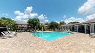 Located at Glen Park in Orange Park, Florida, this outdoor amenity area is designed to provide a welcoming and functional recreational retreat. The centerpiece is a generously sized swimming pool with clear, turquoise water, framed by an expansive paver deck constructed from warm-toned stone pavers that extend across the entire pool surround. The deck offers ample room for sunbathing and relaxation, lined with lounge chairs positioned to take full advantage of the Florida sunshine.

Adjacent to the pool, a covered pavilion serves as the primary gathering and lounge space. The structure features a coffered ceiling supported by clean architectural columns, providing shade and shelter while remaining open to the surrounding landscape. Beneath the pavilion, white-framed outdoor furniture — including cushioned chairs, a loveseat, and a slatted coffee table — creates a comfortable seating arrangement suited for socializing or unwinding.

The area is enclosed by a black wrought iron fence, providing both security and an unobstructed view of the lush greenery and mature trees that border the property. Palm trees and ornamental plantings accent the landscape, reinforcing the warm, Florida-inspired aesthetic. The combination of the pool, spacious paver surround, and shaded pavilion makes this outdoor space a well-rounded community amenity.