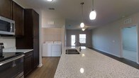This single-story residence features three bedrooms and two bathrooms within a spacious, thoughtfully designed floor plan. The open-concept layout connects the kitchen and living areas seamlessly, creating an inviting atmosphere ideal for both everyday living and entertaining.

The kitchen serves as a central highlight, showcasing a large granite-style island with an undermount sink and elegant pendant lighting overhead. Dark cabinetry provides a striking contrast against the light countertops, while durable wood-look flooring runs throughout the main living areas, adding warmth and cohesion to the space.

The living area, visible from the kitchen, benefits from natural light through rear-facing doors and is equipped with a ceiling fan for year-round comfort. Neutral gray walls throughout contribute to a clean, modern aesthetic that complements a variety of décor styles.

A dedicated laundry area is conveniently located adjacent to the kitchen, complete with a full-size washer and dryer and built-in shelving for additional storage. The open shelving and closet space nearby further enhance the home's practical storage solutions. Overall, the residence combines modern design with functional living across its single level.
