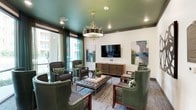 This elegantly designed lounge space makes an immediate impression through its bold, deep green lacquered ceiling — a defining architectural detail that anchors the room's rich color palette. A statement chandelier with gold branch-like accents and a drum shade hangs as a centerpiece, complementing the recessed lighting that illuminates the space throughout.

The seating area is thoughtfully arranged around a tufted leather ottoman-style coffee table, surrounded by a mix of green leather wingback chairs and upholstered patterned armchairs in dark tones. Accent pillows in coordinating botanical prints tie the ensemble together cohesively. A sleek, wall-mounted flat-screen television sits above a mid-century modern slatted wood media credenza, flanked by two large abstract paintings in muted greens and blues that reinforce the overall color story.

Floor-to-ceiling windows dressed in sheer teal drapery line the perimeter, inviting generous amounts of natural light while offering views of the surrounding exterior courtyard. A patterned area rug in grays and blues grounds the space and adds textural depth underfoot.

Additional seating vignettes placed near the windows and walls — accented with glass side tables and bold abstract wall art — provide intimate conversation areas. The overall ambiance is polished yet welcoming, blending contemporary design with warm, curated details.