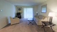 This single-floor space centers around a welcoming living room designed for both comfort and functionality. The room is finished with wall-to-wall carpeting in a neutral tone, contributing to a warm and relaxed atmosphere throughout. Soft, light-colored walls enhance the brightness of the space, creating an open and airy feel.

A large sliding glass door fitted with vertical blinds serves as the focal point of the room, allowing abundant natural light to fill the interior while providing direct access to an outdoor area. The living room accommodates a generously sized sofa and a matching armchair, both upholstered in a light cream fabric, arranged around a dark-framed X-base coffee table that adds a contemporary contrast to the soft furnishings.

Additional seating and accent tables are positioned throughout the room, offering practical layout flexibility. A television setup occupies one corner, accompanied by a low media console, while framed wall art and a floor lamp contribute decorative touches. The overall ambiance is clean, understated, and well-lit, making the space feel inviting and move-in ready.