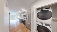 Located on Harvard Avenue in Seattle's vibrant Capitol Hill neighborhood, this efficiently designed single-floor unit offers a thoughtfully laid-out living space with modern finishes throughout. The galley-style kitchen is a standout feature, boasting white upper cabinetry with matte black hardware, two-toned dark lower cabinets, crisp white quartz countertops, and a light tile backsplash. A full suite of stainless steel appliances includes a French door refrigerator, electric range, over-the-range microwave, and dishwasher, making the kitchen both stylish and fully functional. Warm-toned hardwood flooring runs throughout the main living areas, complementing the clean white walls and recessed lighting that keep the space feeling bright and airy. A dedicated in-unit laundry area houses a stacked GE front-load washer and dryer, along with a built-in shelf for added storage convenience. The bathroom features cool-toned concrete-style tile flooring, a generous vanity with white countertop and matte black hardware, a large mirror, and clean white walls that create a crisp, minimal aesthetic. A hallway connects the key areas of the unit, contributing to a logical and efficient flow throughout the space.