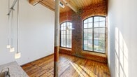 This two-story loft apartment at 1101 Hawthorne Lane blends industrial character with modern finishes. The main level features a dramatic double-height kitchen with pendant lighting, granite countertops, a large island with a stainless steel sink, rich dark cabinetry, and stainless steel appliances. Original hardwood floors and structural columns run throughout. A stunning living area showcases exposed brick walls, soaring arched windows that flood the space with natural light, and a wood-planked ceiling. The upper level hosts the spacious primary bedroom with exposed beams and industrial-style ceilings. Both bathrooms are finished with granite vanities and dark wood cabinetry, with one featuring a full tub-shower combo.