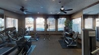 This fitness center is a thoughtfully designed, single-floor exercise facility that offers a comprehensive workout environment. The space is outfitted with a wide variety of equipment, including treadmills, upright and recumbent stationary bikes, elliptical machines, and multi-function strength training stations, providing options for both cardiovascular conditioning and resistance training.

The interior features a neutral-toned, textured carpet that defines workout zones, complemented by dark rubber equipment mats. Wood-trimmed crown molding and rustic-style door frames lend a warm, refined aesthetic to the space, while a textured ceiling adds visual depth. Multiple ceiling fans are evenly distributed throughout the room, ensuring consistent air circulation during workouts.

Large windows line the perimeter walls, inviting abundant natural light and offering views of the surrounding outdoor environment. Horizontal blinds allow for adjustable light control. Full-length mirrors on one wall visually expand the space and support proper exercise form. A decorative potted tree adds a natural element to the corner of the room.

Recessed lighting supplements natural light for well-illuminated workouts at any time of day. A set of wood-framed glass doors provides access to an adjacent area, further enhancing the open, connected feel of this well-appointed fitness facility.