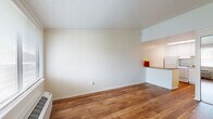 This single-floor unit offers a cohesive, efficient layout with warm-toned hardwood flooring running throughout the main living areas, creating a sense of continuity and warmth. The living area is open and bright, featuring neutral white walls, textured ceilings with crown molding, and natural light filtering in through a window near the entryway. The space flows naturally toward the kitchen, which is separated by a peninsula counter featuring a stone-look surface — ideal for casual dining or meal prep. The kitchen is well-appointed with white cabinetry extending to the ceiling, a skylight for added brightness, a white refrigerator, and a white range with overhead storage. A utility area is accessible from the kitchen. The bathroom includes a white vanity with under-cabinet storage, brushed nickel fixtures, a wide mirror, and chrome towel rings. Consistent hardwood flooring ties the bathroom into the rest of the unit seamlessly. The layout, as seen in the 3D overview, is thoughtfully arranged to maximize usability within a compact footprint, making it a practical and tidy living space.