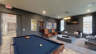 Located at 200 W Main St in New Albany, Ohio, this single-story community clubhouse is designed with a refined, contemporary aesthetic that balances comfort and sophistication. The space is divided into two distinct yet complementary zones. The lounge and recreation area is anchored by deep charcoal-gray walls, a plush gray sofa, and a pair of rich cognac leather wingback chairs arranged around a substantial concrete-style coffee table. A wall-mounted flat-screen TV and a clustered gold pendant light fixture add a modern, curated feel, while a full-size billiards table serves as a central social focal point. Framed black-and-white artwork lines the walls, and natural light filters in through tall, grid-paned windows. A bar-height seating area provides an additional gathering option within this wing. The adjoining dining and event room offers a notably different atmosphere, with bright white walls, intricate herringbone-patterned wood-look flooring, and elegant round white tables paired with black velvet and gold-accented chairs. A decorative display shelf and pendant chandelier enhance the room's upscale ambiance. French doors connect the two areas, making the overall space highly adaptable for both casual socializing and more formal gatherings.